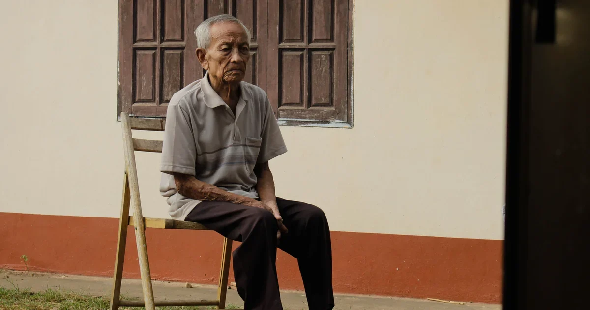 An elderly man sitting on a wooden chair outside his home, looking into the distance