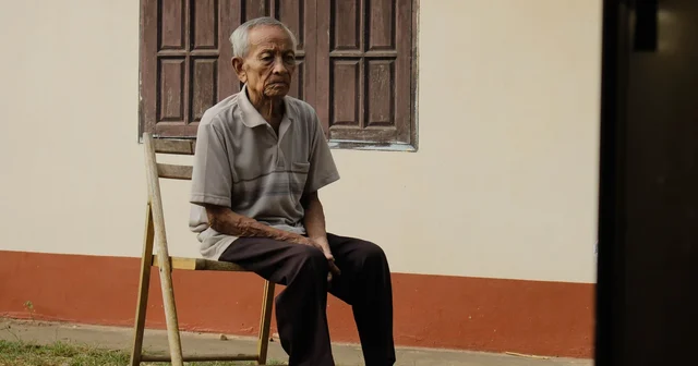 An elderly man sitting on a wooden chair outside his home, looking into the distance