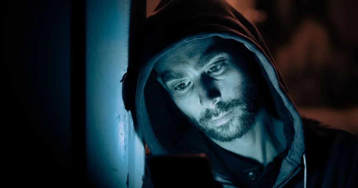 A bearded man in a dark hoodie looking at a smartphone, his face lit by the screen