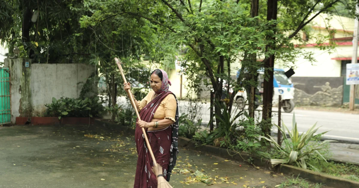 A woman in traditional attire sweeping outdoors in a green area of Raipur, India
