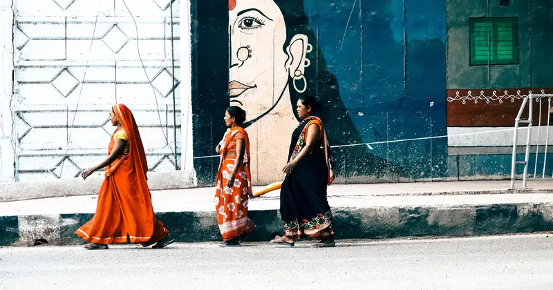Three women in saris walking together past a colourful mural on a city pavement in India