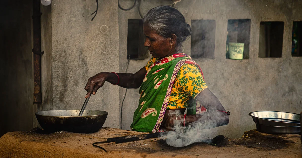 A rural Indian woman in bright clothing prepares food outdoors, a daily unpaid task for millions of women